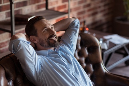 Feeling great. Positive happy bearded businessman sitting on the sofa and putting hands behind his head while enjoying his relaxationの写真素材