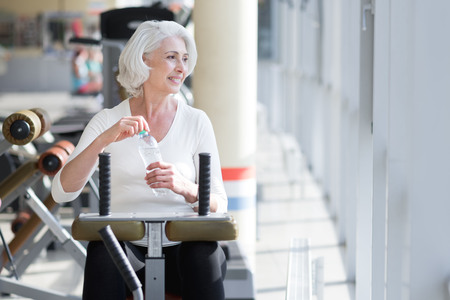 Pleasant relaxation. Charming attractive senior woman drinking water and relaxing after working out in the gym.の写真素材