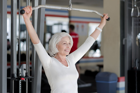 Inspiring fitness. Joyful athletic senior woman smiling and training back muscles by using sports...
