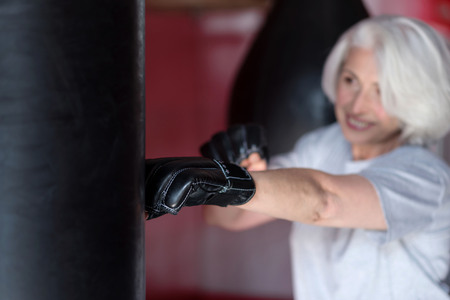 Just box. Cheerful pretty senior woman boxing punching bag in a gym and smilingの写真素材