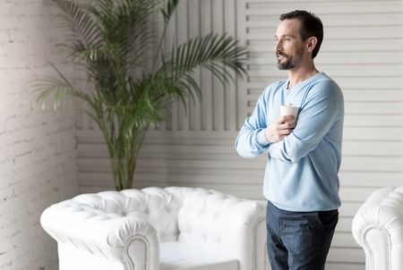 Joy and happiness. Nice cheerful good looking man crossing his arms and holding a cup while standing in the roomの写真素材