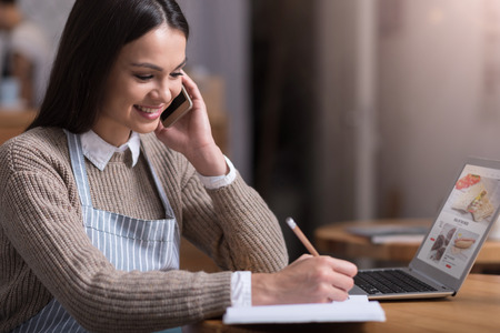 Lovely work. Cheerful beautiful young woman smiling and making notes while talking on cellphone.の写真素材