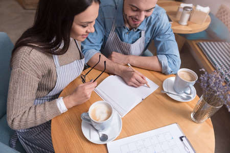 Coffee pause. Positive young charming couple of cafe owners drinking tea and smiling while working by the table.の写真素材