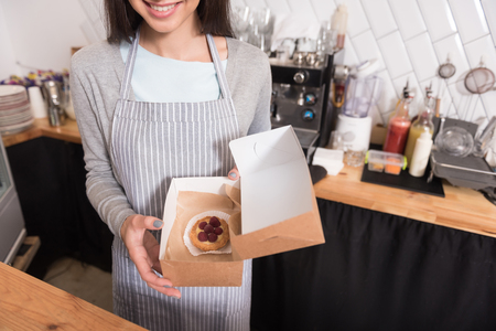 Service with a smile. Charming joyful young waitress smiling and presenting cake in a box while working at the counter in a cafe.の写真素材