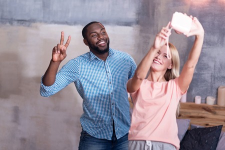 Funny moments. International young pretty couple making selfie together while laughing and standing in bedroom.の写真素材