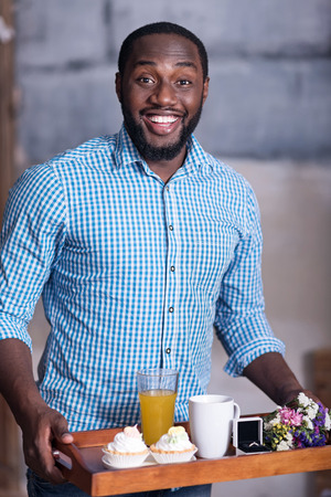 It is coming. African young overjoyed man preparing for marriage proposal while holding a tray with breakfast and the ring and smiling.の写真素材
