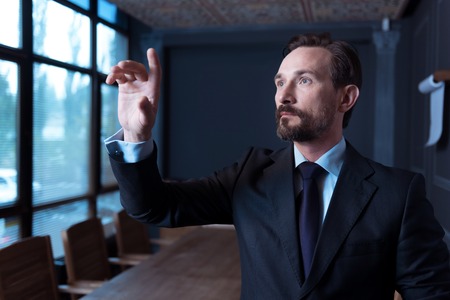 Age of technology. Good looking confident bearded man standing in the conference hall and touching the virtual sensory panel while working on itの写真素材