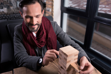 Way to relax. Delighted good looking joyful man removing one block from the tower and concentrating on it while playing blocksの写真素材