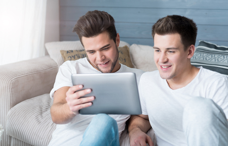 Pleasure for two. Happy cute gay couple sitting on the flor and smiling while using tablet.の写真素材