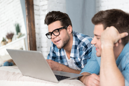 Pleasant relaxation together. Cheerful young handsome men smiling and using a laptop while relaxing together on a bed.の写真素材