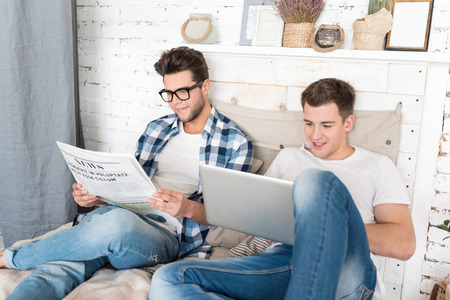 Leisure time. Happy young gay couple reading together while holding newspaper and using laptop in...