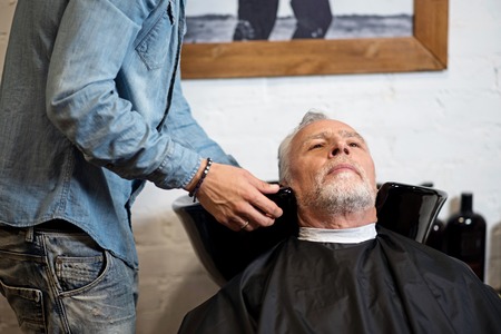 Waiting for refreshment. Thoughtful aged man sitting in barber shop while his hair being washed by specialist.の写真素材