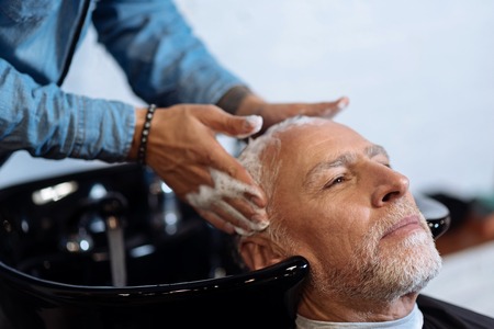 Perfect massaging. Close-up portrait of pleasant bearded man having his hair washed by hairdresser in barber shop.の写真素材