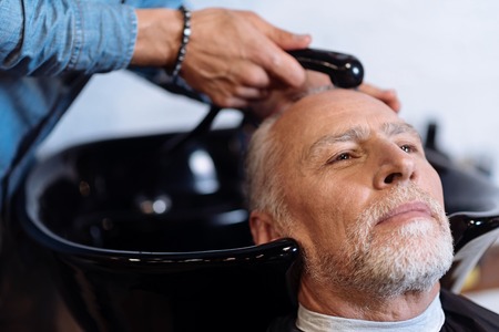Enjoying service. Close up portrait of senior bearded man sitting in barber shop while professional...
