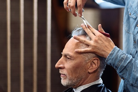 Jewelry work. Side view of pleasant senior man sitting in barbershop while his grey hair being cut by professional.の写真素材