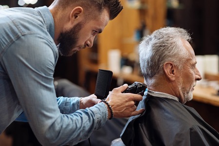 Just little bit. Side view, young handsome hairdresser trimming neck of sitting senior client with grey hair at barbershop.の写真素材