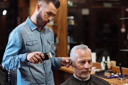 Detailed work. Young handsome hairdresser standing and accurately trimming hair of old barbershop client.の写真素材