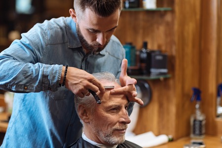 Without any words. Attractive young barber standing and cutting forehead hair on senior client by using special knife at barbershop.の写真素材