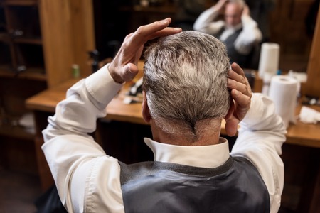 Beautify yourself. Close up back view of senior gentle man styling his grey hair himself at barbershop.の写真素材