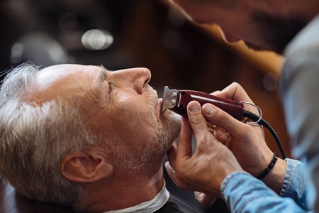 Small details. Close up portrait of senior man with grey hair getting his beard shaved by professional hairdresser at barbershop.の写真素材