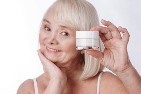 Facial care. Happy cheerful nice woman holding a cream bottle and smiling while using cosmeticsの写真素材