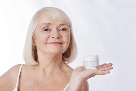 Beauty products. Positive smiling aged woman having a cream bottle on her palm and smiling while presenting beauty productsの写真素材
