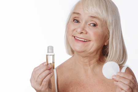 Cosmetic products. Elated happy elderly woman holding a cleansing lotion and a cotton pad and smiling while using cosmetic productsの写真素材