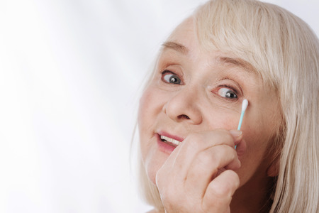 Cotton swabs. Delighted positive aged woman holding a cotton swab and using it while caring about her appearanceの写真素材
