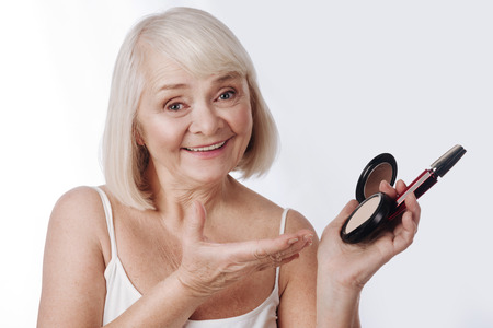 Stay beautiful. Nice pleasant retired woman holding face powder and mascara and pointing at it while looking at youの写真素材