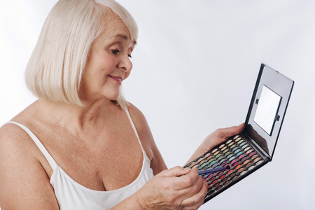 Wide choice of colour. Good looking cheerful senior woman holding a makeup brush and choosing the color while applying eyeshadowの写真素材