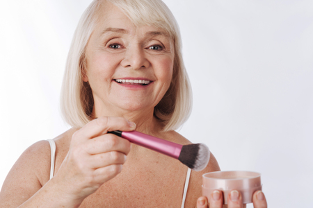 Reducing skin shine. Good looking happy retired woman holding a makeup brush and using it while applying face powderの写真素材