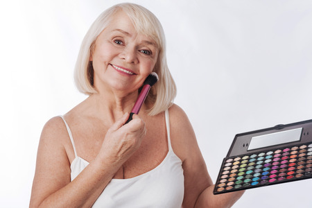 Professional make up. Joyful grey haired retired woman holding an eyeshadow palette and using a cosmetic brush while putting on professional make upの写真素材