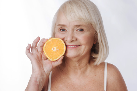Citrus fruits. Pleasant joyful content woman holding an orange half and squeezing it while looking at youの写真素材