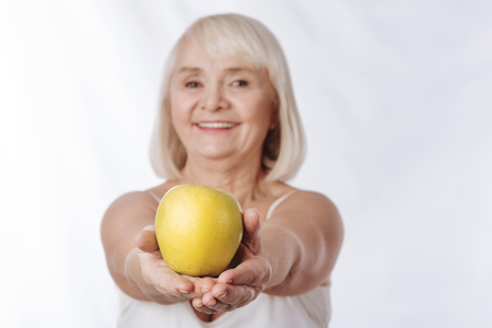Full of vitamins. Selective focus of a big yellow apple being in hands of a positive charming smiling woman while being offered to youの写真素材