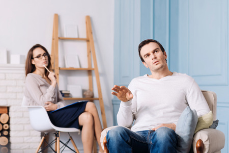 Reveal your thoughts. Pleasanttal talkative handsome man sitting in the armchair and discussing his problems while professional psychologist sitting in the backgroundの写真素材