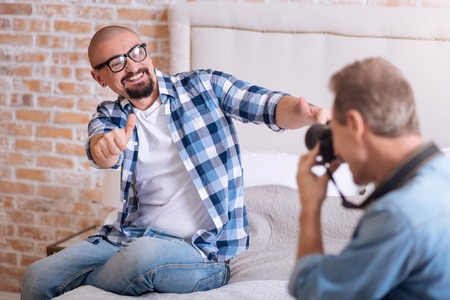 Full of delight. Delighted joyful cheerful homosexual man sitting on the bed and posing while smiling and his partner taking the picture of himの写真素材