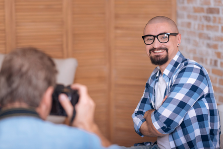Smiling for a portrait. Happy joyful cheerful man sitting in the studio and posing while being with crossed arms and the photographer taking the picture of himの写真素材