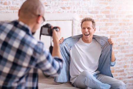 Full of joy . Delighted joyful happy homosexual man sitting on the bed and posing while smiling and his partner taking the picture of himの写真素材