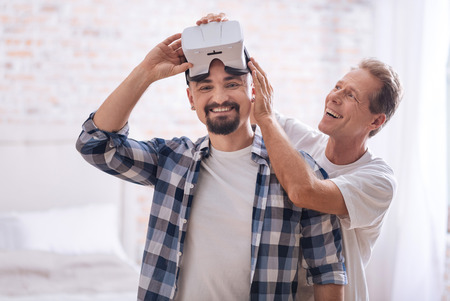 We spending enjoyably our free time. Smiling cheerful delighted non-traditional couple standing at home and testing virtual reality glasses while expressing happiness and interestの写真素材