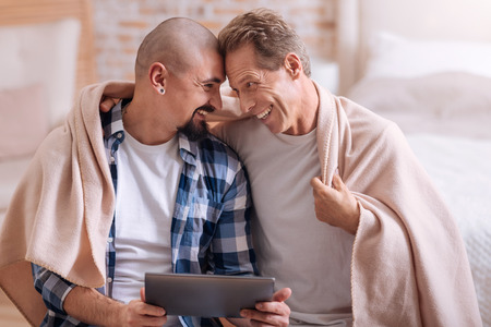 So cute to be with you. Smiling happy positive non-traditional couple sitting in the bedroom and hugging while using the tablet and being covered with the blanketの写真素材