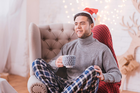 New Year time. Handsome nice happy man sitting in a chair and holding a tea cup while wearing a Santa hatの写真素材