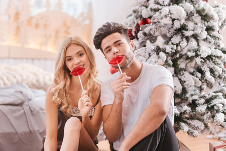 Eating lollipops. Positive delighted happy couple sitting under the Christmas tree and holding lollipops while having fun togetherの写真素材