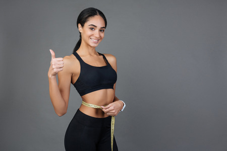 Good results. Positive delighted pleased sportswoman wearing black sport suit keeping her right hand bent in elbow with thumb up while measuring waistの写真素材