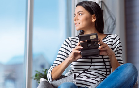 Full of inspiration. Inspired thoughtful young woman sitting on the chair at home and holding the camera while expressing inspiration and looking at the windowの写真素材