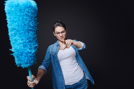 You will be the next. Delighted determined young woman holding a dusting brush and standing against black background while smiling and pointingの写真素材