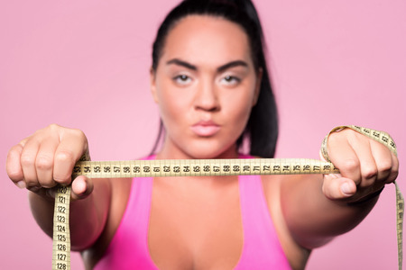 My aim. Close up portrait of serious mulatto woman holding body measuring tape against isolated pink...