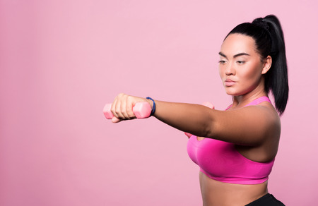 Getting stronger. Pretty chubby mulatto woman doing boxing exercises with dumbbells while standing against isolated pink background.の写真素材