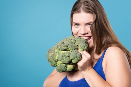 Low carb. Portrait of happy smiling chubby girl biting off broccoli and eating it while standing isolated on blue background.の写真素材