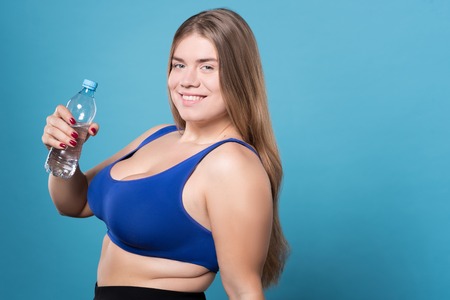 Water is life. Close up of nice young smiling chubby woman standing against isolated blue background with bottle of water.の写真素材