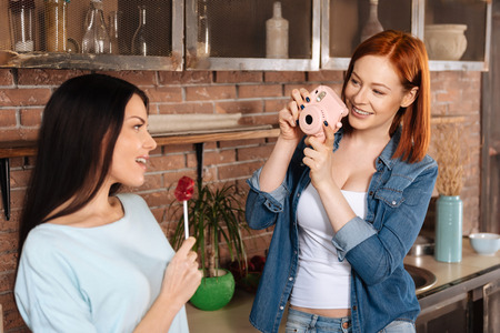 Look at me. Positive delighted women wearing casual clothes holding photo camera in front of face while shooting her friendの写真素材
